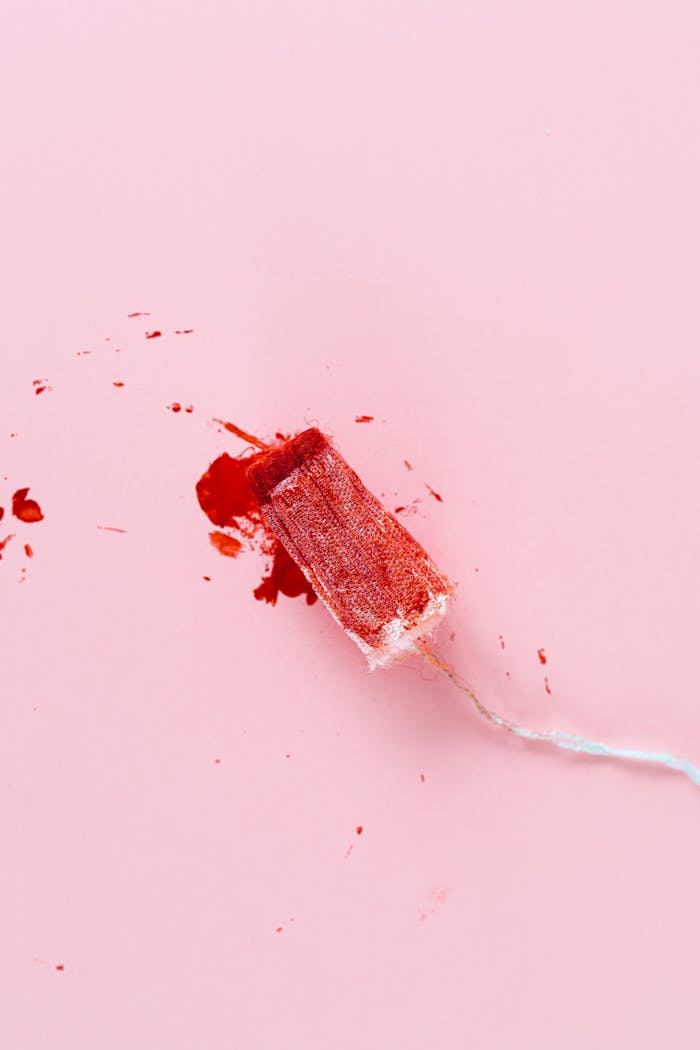 services-02 A high-angle view of a tampon with red stains on a soft pink background, symbolizing menstruation.